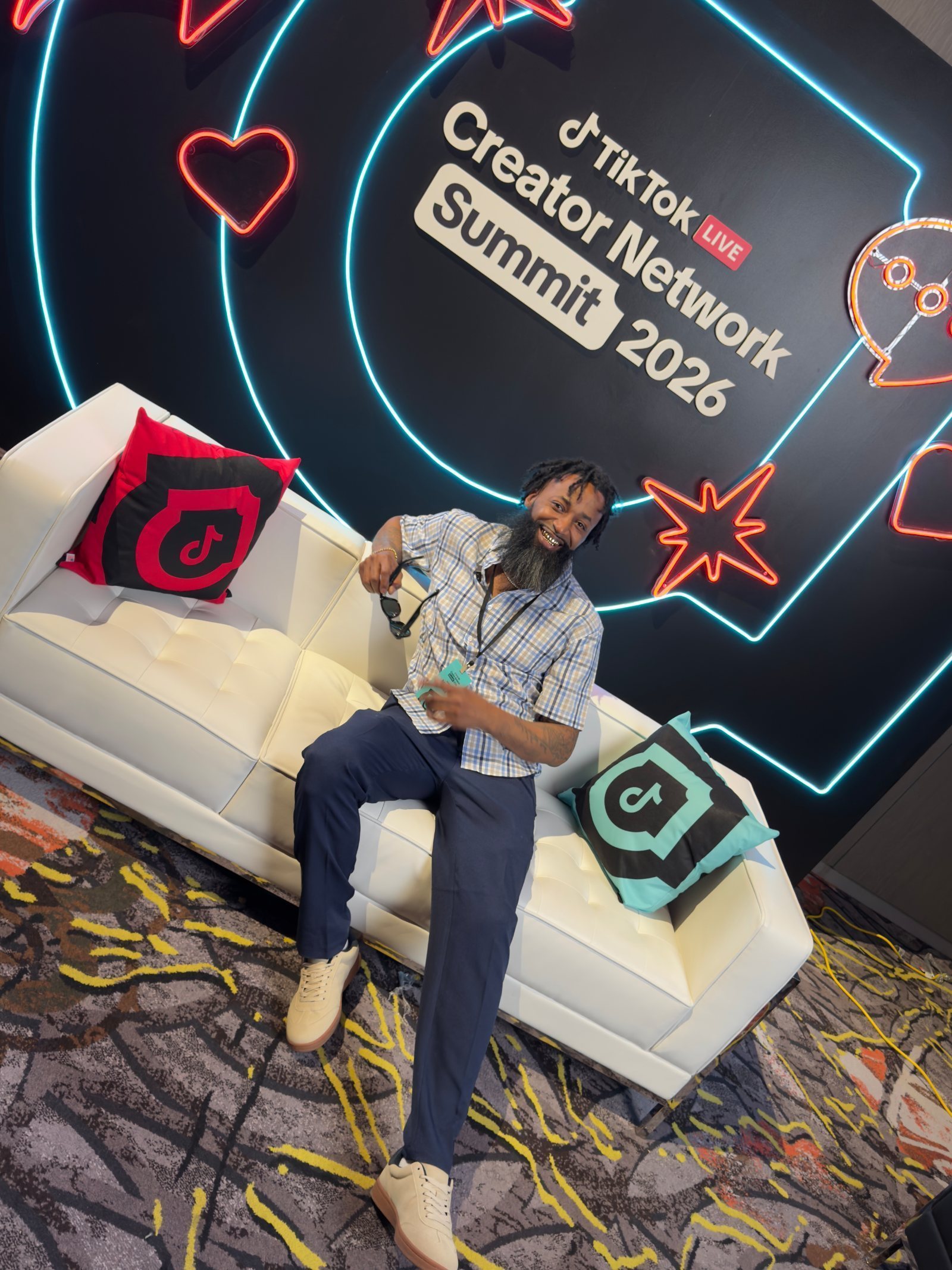 Whoa Nahh seated on the white tufted couch at the creator-network summit with the branded pillows and neon signage behind him